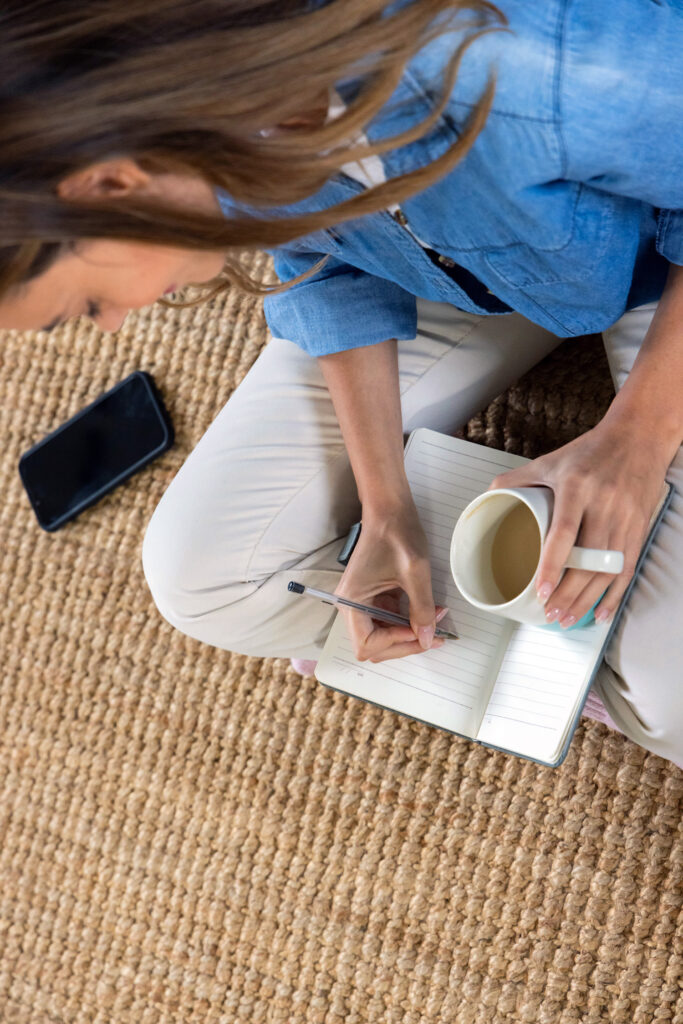 Writing in notebook, woman holding coffee cup, sitting on woven rug at home. Lifestyle, cozy, relaxation, morning, beverage, casual